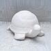 Handmade unpainted ceramic turtle on a lacy ecru table cloth and a curtain behind it.  It is facing slightly toward the left.